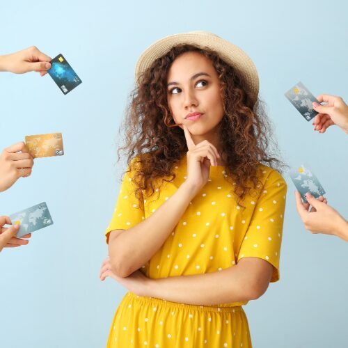 Young woman in a yellow polka-dot dress and hat, thinking as several hands offer her different credit cards against a blue background.